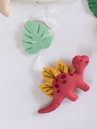 Handcrafted felt Stegosaurus from the Roar Away baby cot mobile with tropical leaf accents, adding playful prehistoric charm to nursery décor.
