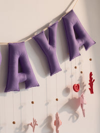 Close-up of the handcrafted Miraya purple felt letter banner featuring hanging felt sea creatures, coral shapes, and wooden beads on a cream fabric string.