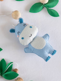 Cute blue handcrafted felt hippopotamus from the Baby Being cot mobile, beautifully paired with wooden beads and green felt leaves for safari nursery styling.