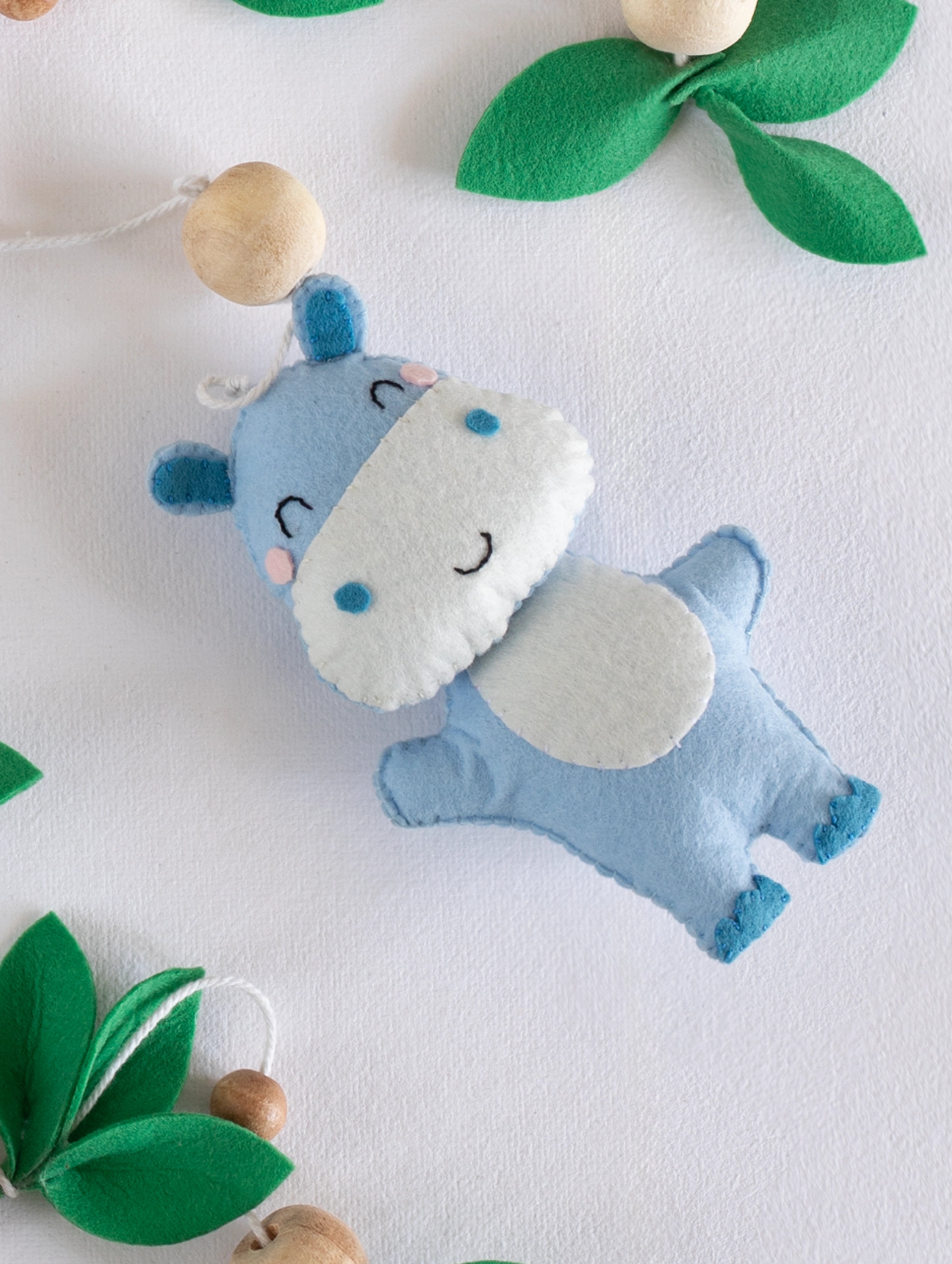 Cute blue handcrafted felt hippopotamus from the Baby Being cot mobile, beautifully paired with wooden beads and green felt leaves for safari nursery styling.