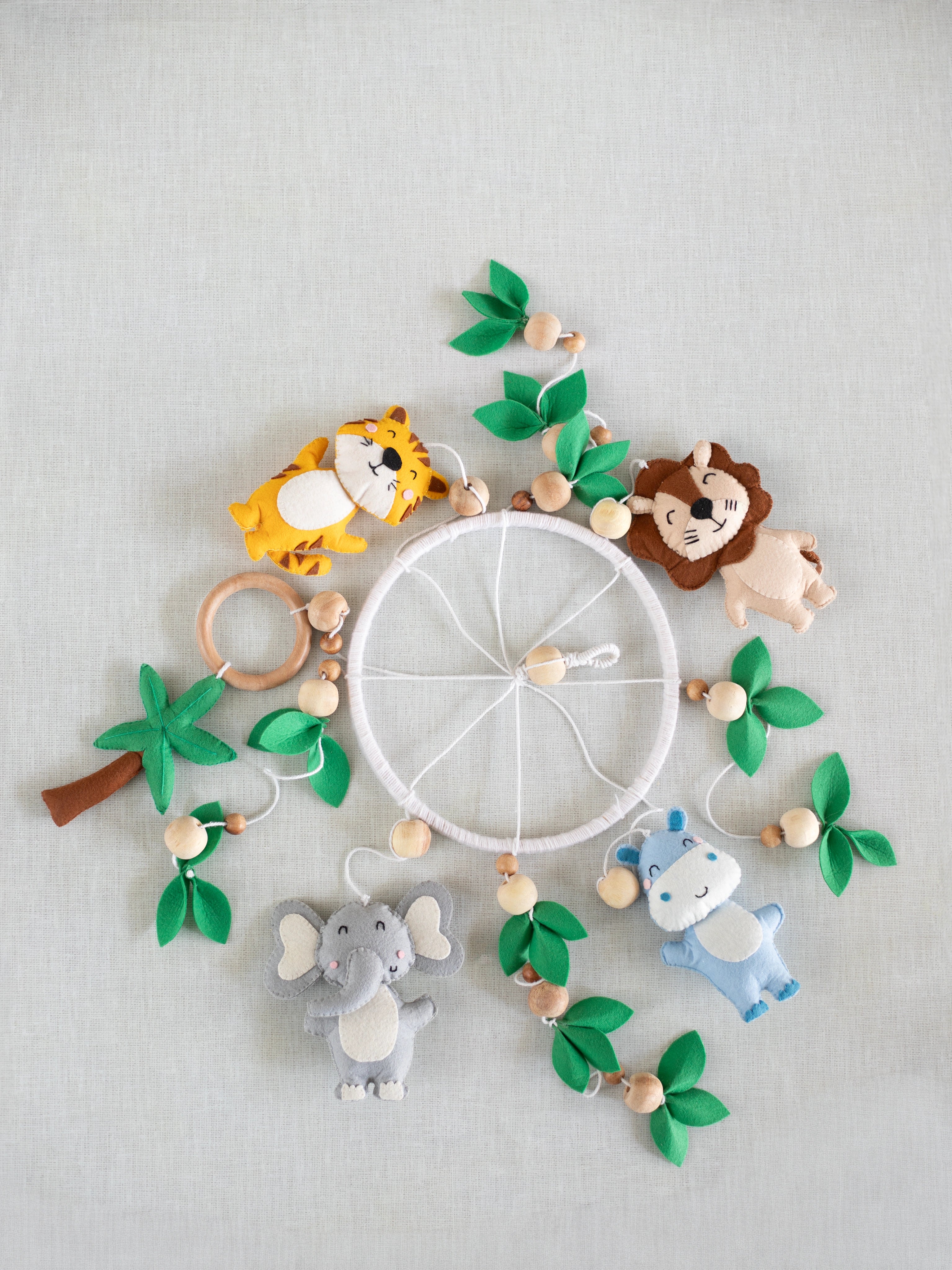 Baby Being handcrafted felt cot mobile featuring a lion, tiger, elephant, hippo, palm tree, wooden teether ring, and leafy details for a safari-inspired nursery.