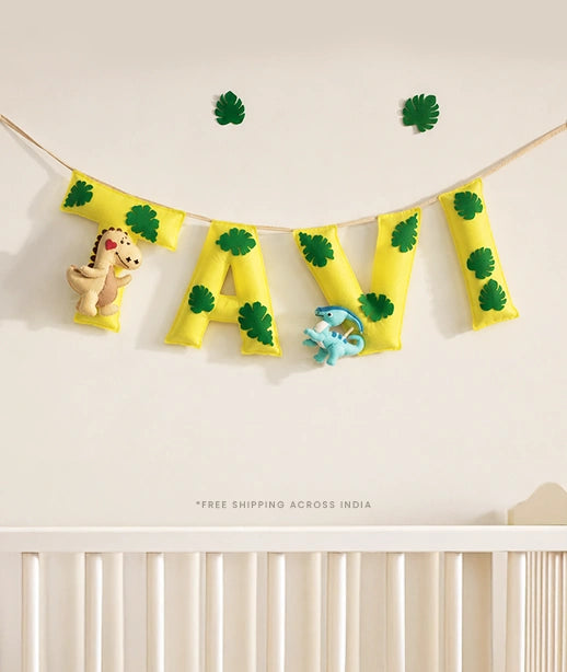 Custom handmade yellow felt name banner spelling TAVI with green leaf prints and dinosaur plush toys, displayed above a wooden baby play gym with hanging animal  — personalized nursery wall decor and baby shower gift, free shipping across India