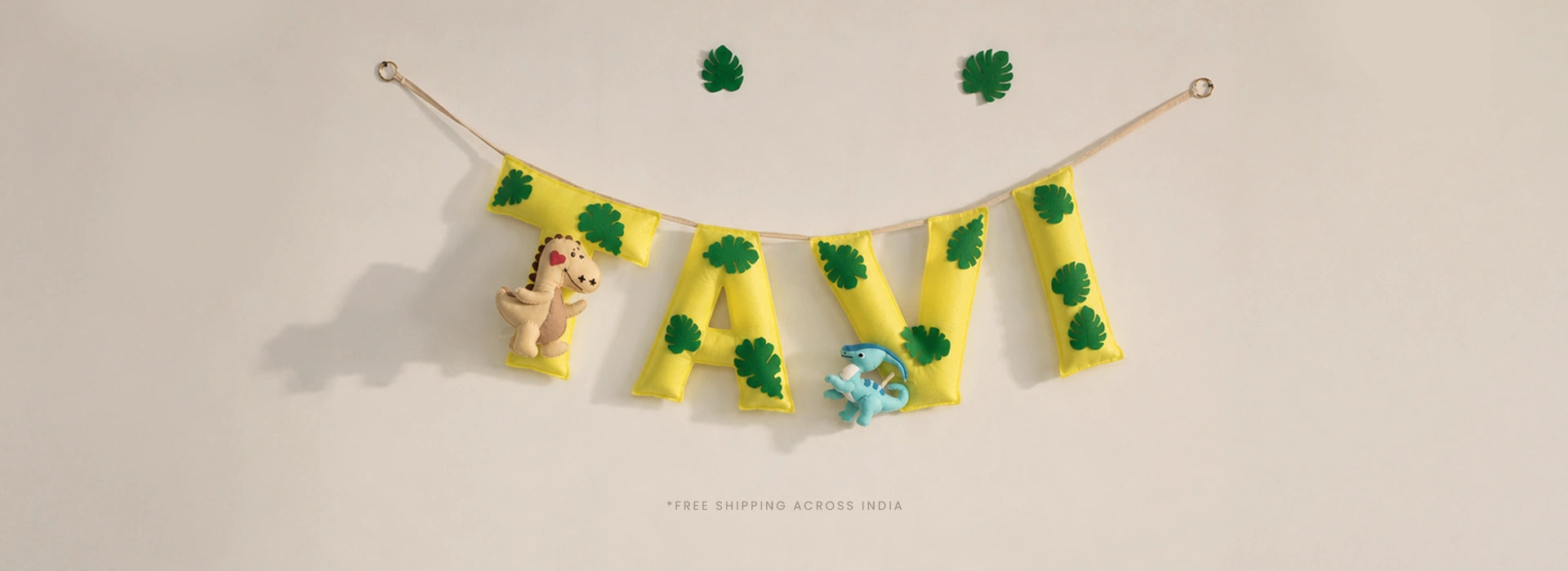 Custom handmade yellow felt name banner spelling TAVI with green monstera leaf prints and dinosaur plush charms, hung on a rope string — personalized nursery wall decor and baby shower gift, free shipping across India