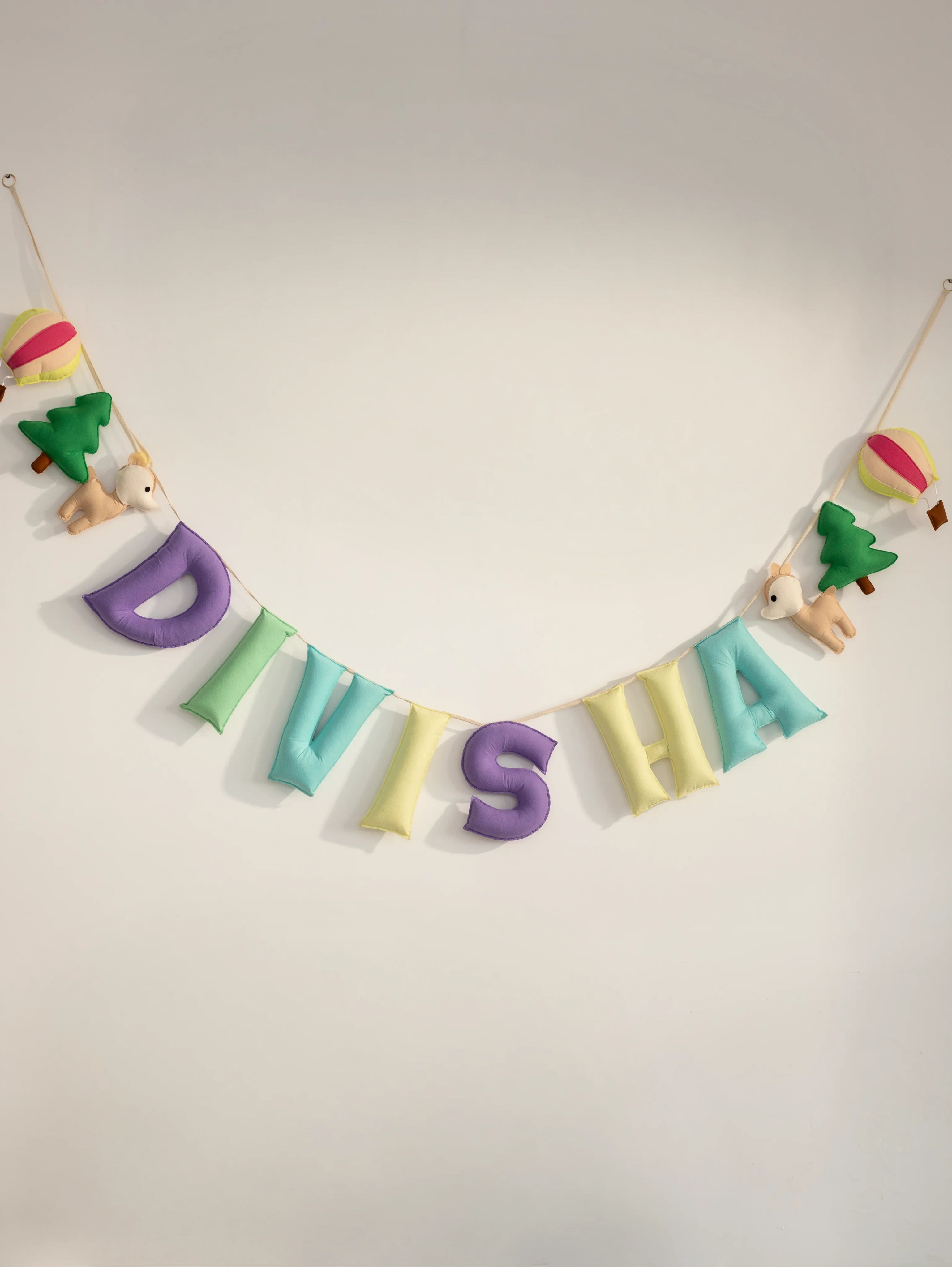 Full view of a personalized handmade felt name banner spelling "DIVISHA" with soft pastel letters, woodland deer, pine trees, and hot air balloons, ideal for baby nursery wall decoration.