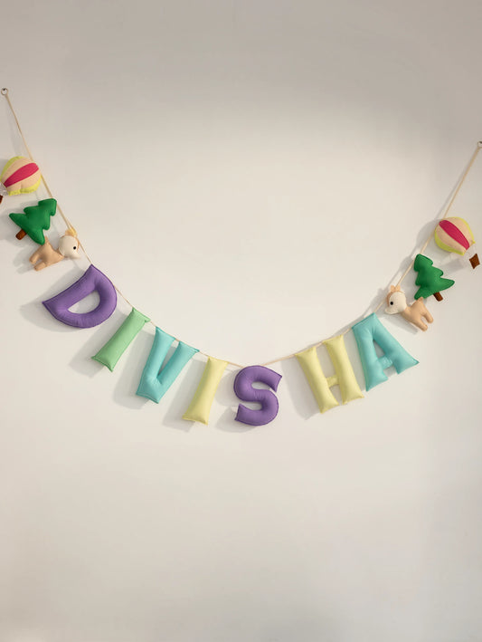 Full view of a personalized handmade felt name banner spelling "DIVISHA" with soft pastel letters, woodland deer, pine trees, and hot air balloons, ideal for baby nursery wall decoration.