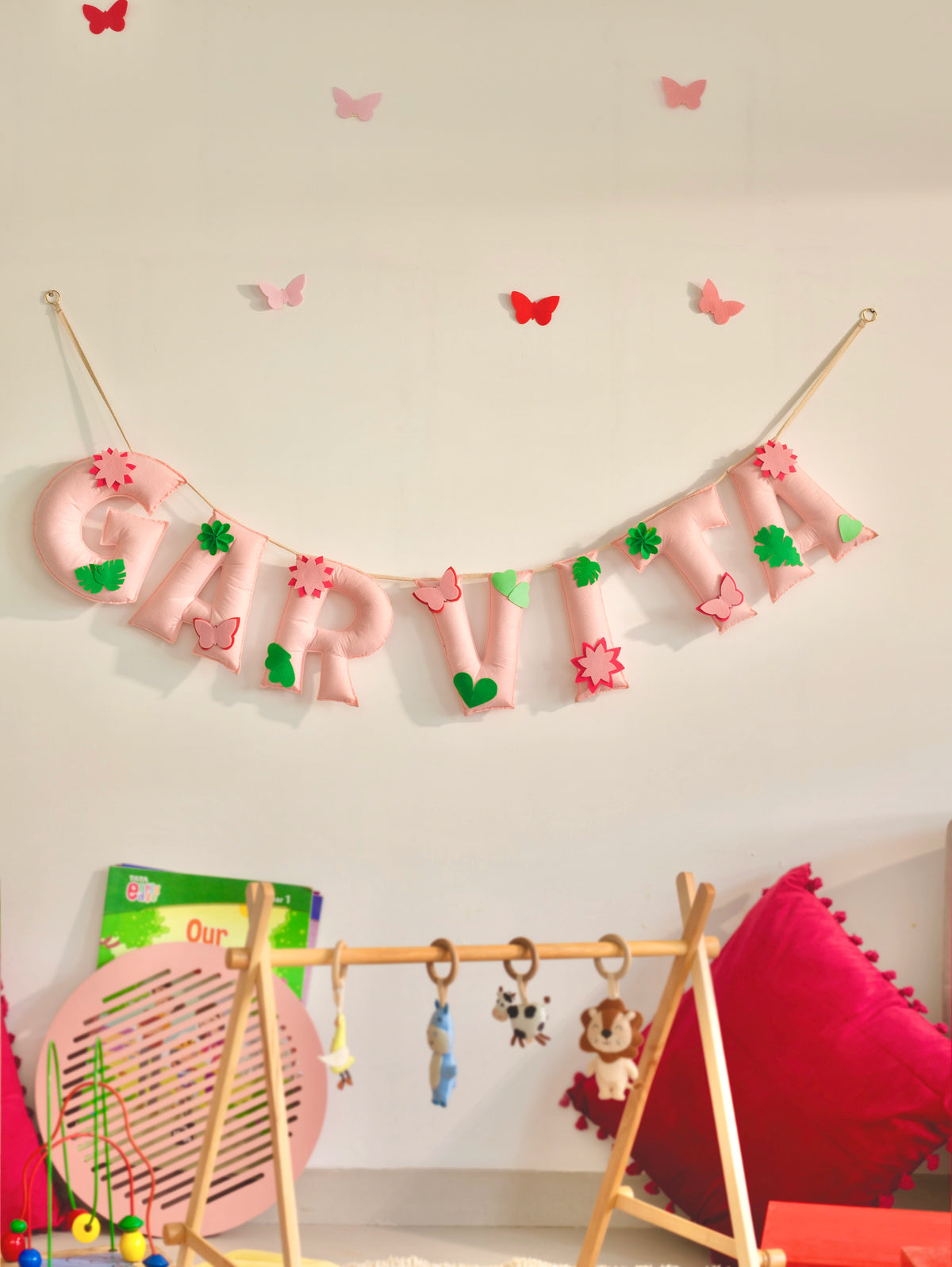 Close-up of a soft pink Blossom Banner for kids’ nursery décor, featuring padded fabric letters decorated with layered pink felt flowers, green tropical leaves and floral accents, suspended on a natural jute string against a neutral wall.