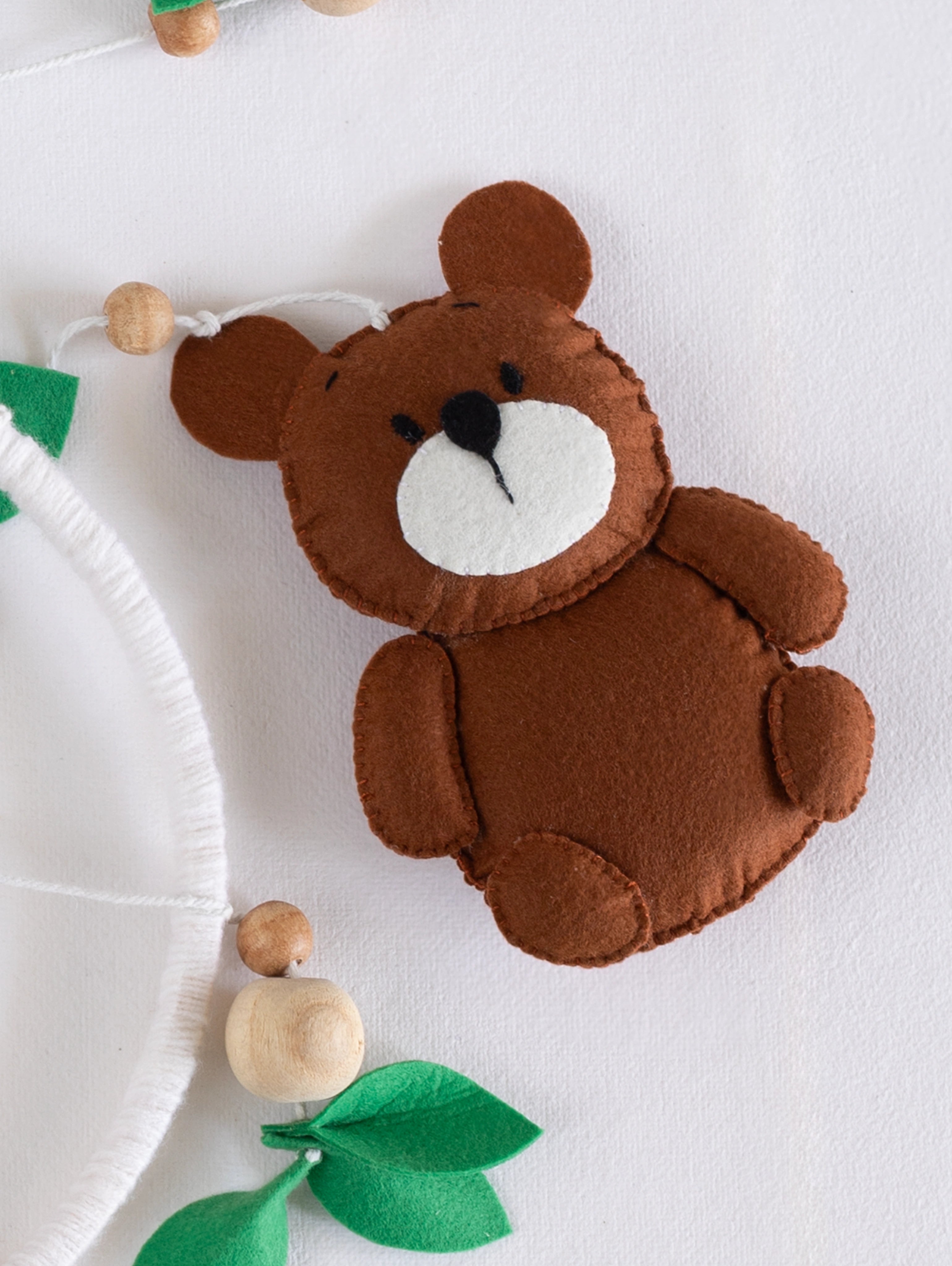 Adorable handcrafted felt bear ornament from the Into the Wild baby mobile, accented with natural wooden beads and forest greenery for nursery styling.