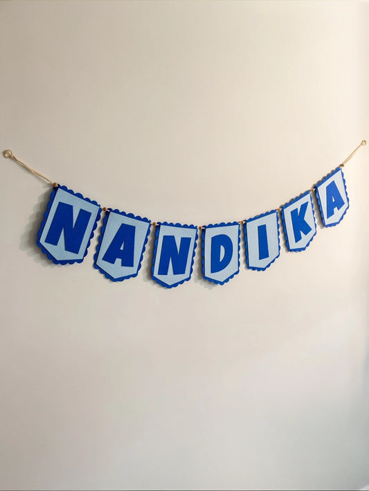 Personalized blue felt name banner spelling "Nandika" hanging on a nursery wall with handcrafted alphabet flags.