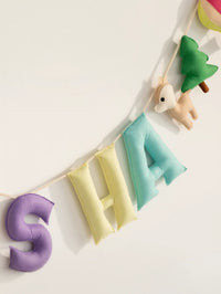 Close-up of a handmade personalized felt name banner featuring soft pastel letters "SHA" with woodland-themed decorations including a deer, tree, and hot air balloon for baby nursery decor.