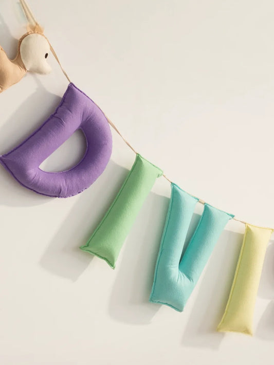 Close-up of a personalized handmade felt name banner displaying pastel letters "DIVI" suspended on a cotton string, perfect for baby nursery wall decor and personalized gifting.