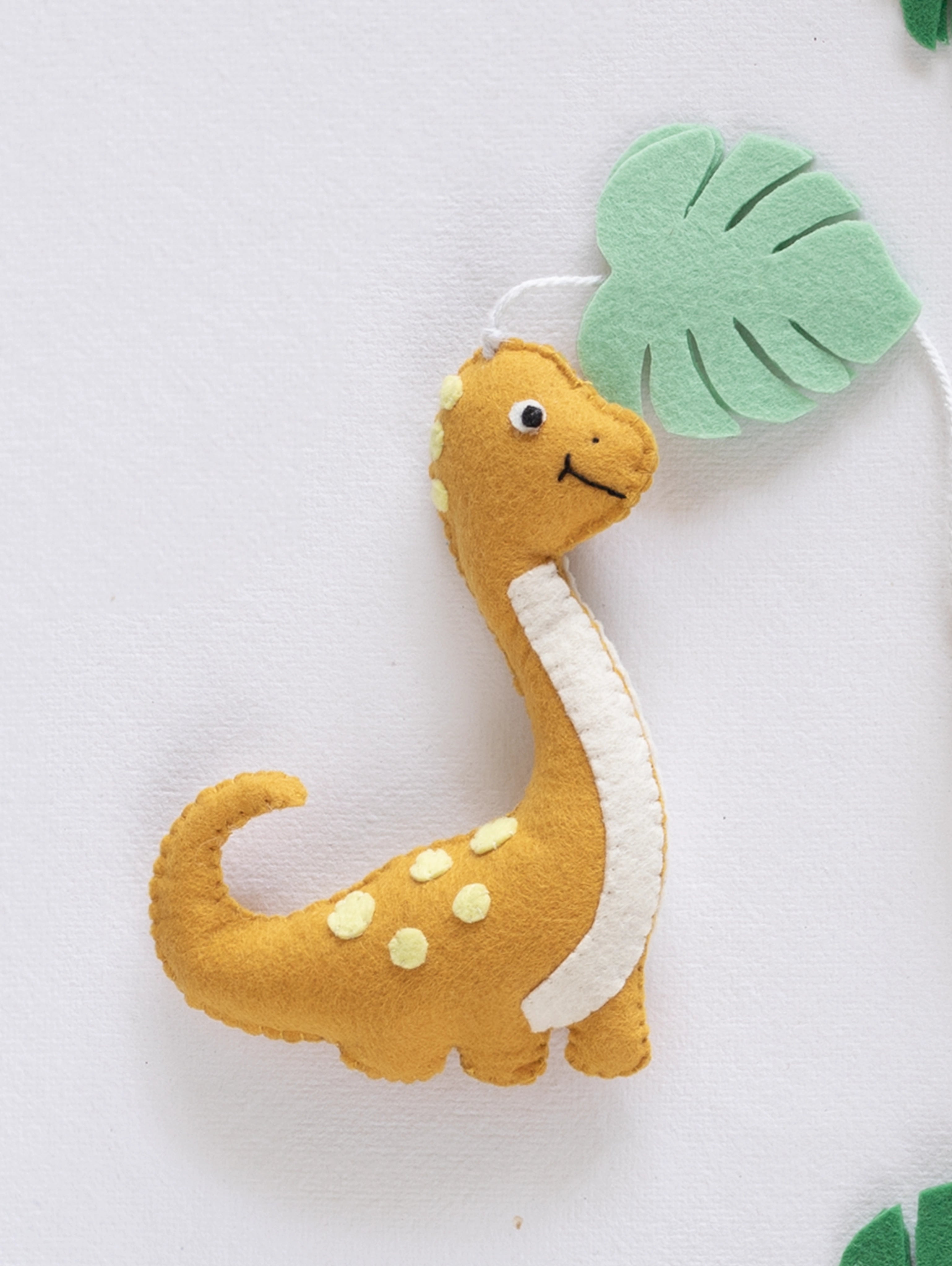 Pastel handcrafted Brontosaurus from the Roar Away baby mobile featuring jungle-inspired felt leaves for a cozy dinosaur nursery theme.