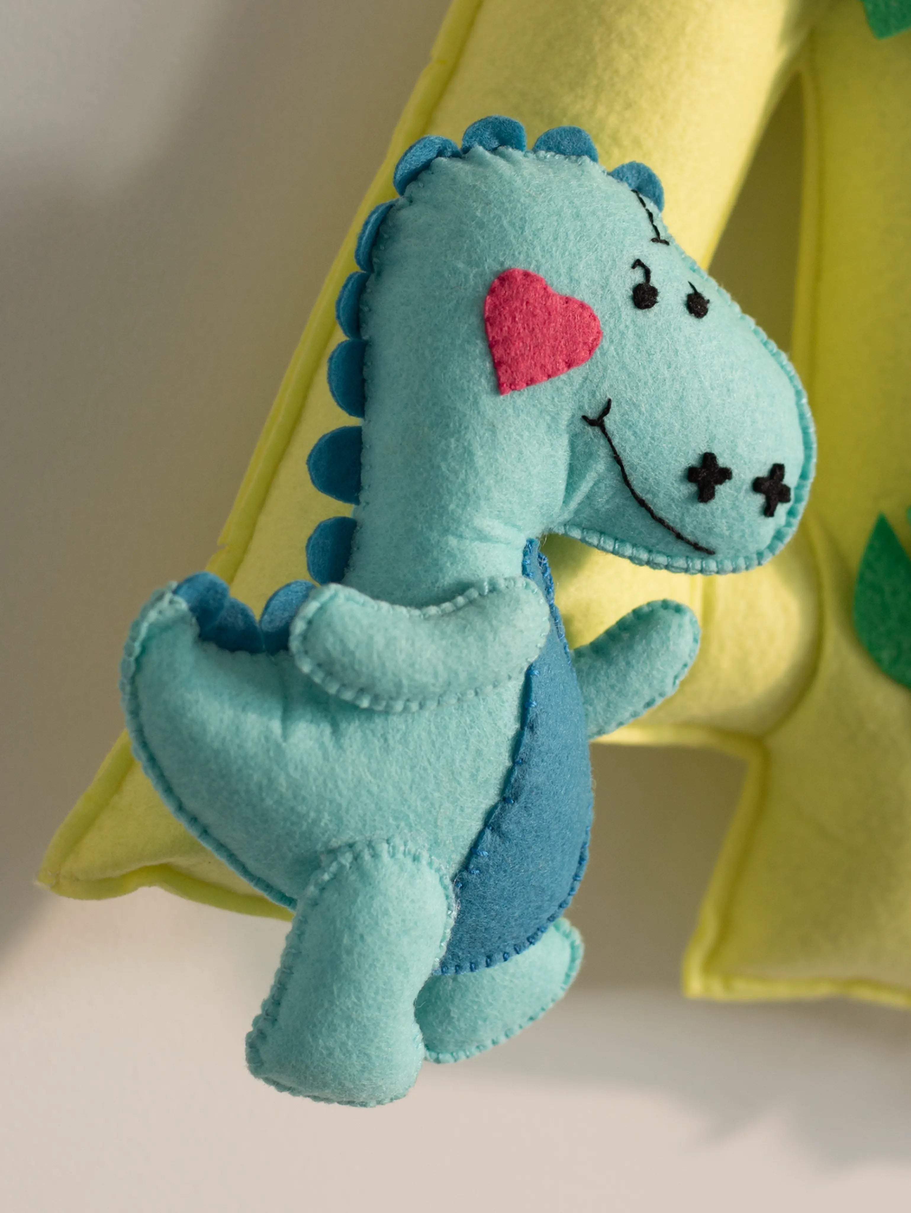 Close-up of a handcrafted blue felt dinosaur ornament attached to the personalized Arav nursery name banner