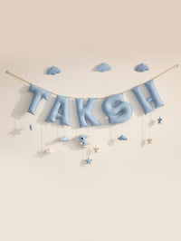A pastel blue letter banner spelling 'TAKSH' hangs from a string against a warm beige wall, decorated with small blue clouds, stars, and a koala with moon shaped ornament.