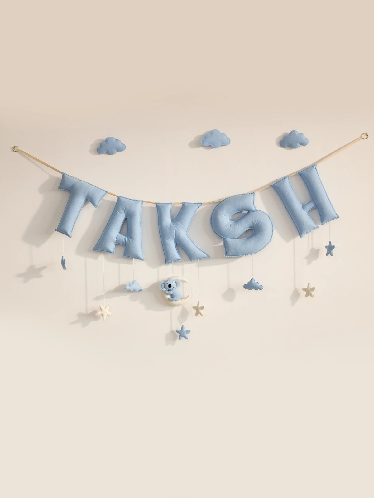 A pastel blue letter banner spelling 'TAKSH' hangs from a string against a warm beige wall, decorated with small blue clouds, stars, and a koala with moon shaped ornament.
