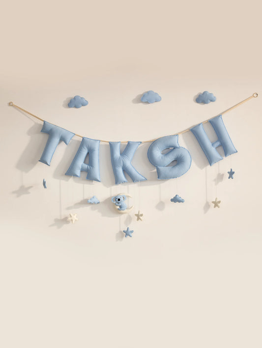 A pastel blue letter banner spelling 'TAKSH' hangs from a string against a warm beige wall, decorated with small blue clouds, stars, and a koala with moon shaped ornament.