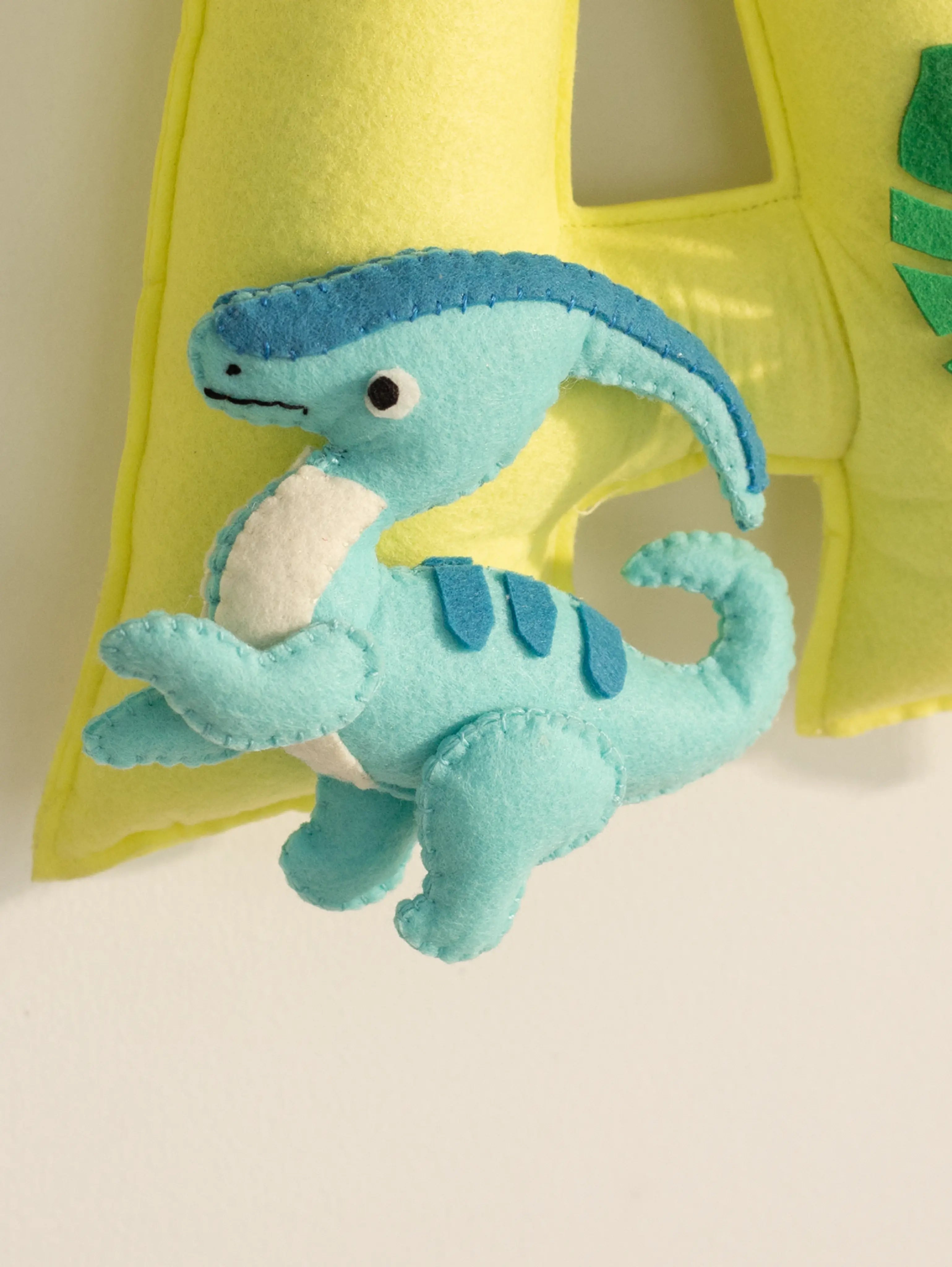 Close-up of a blue felt dinosaur beside the letter ‘A’ on the personalized Arav baby name banner.