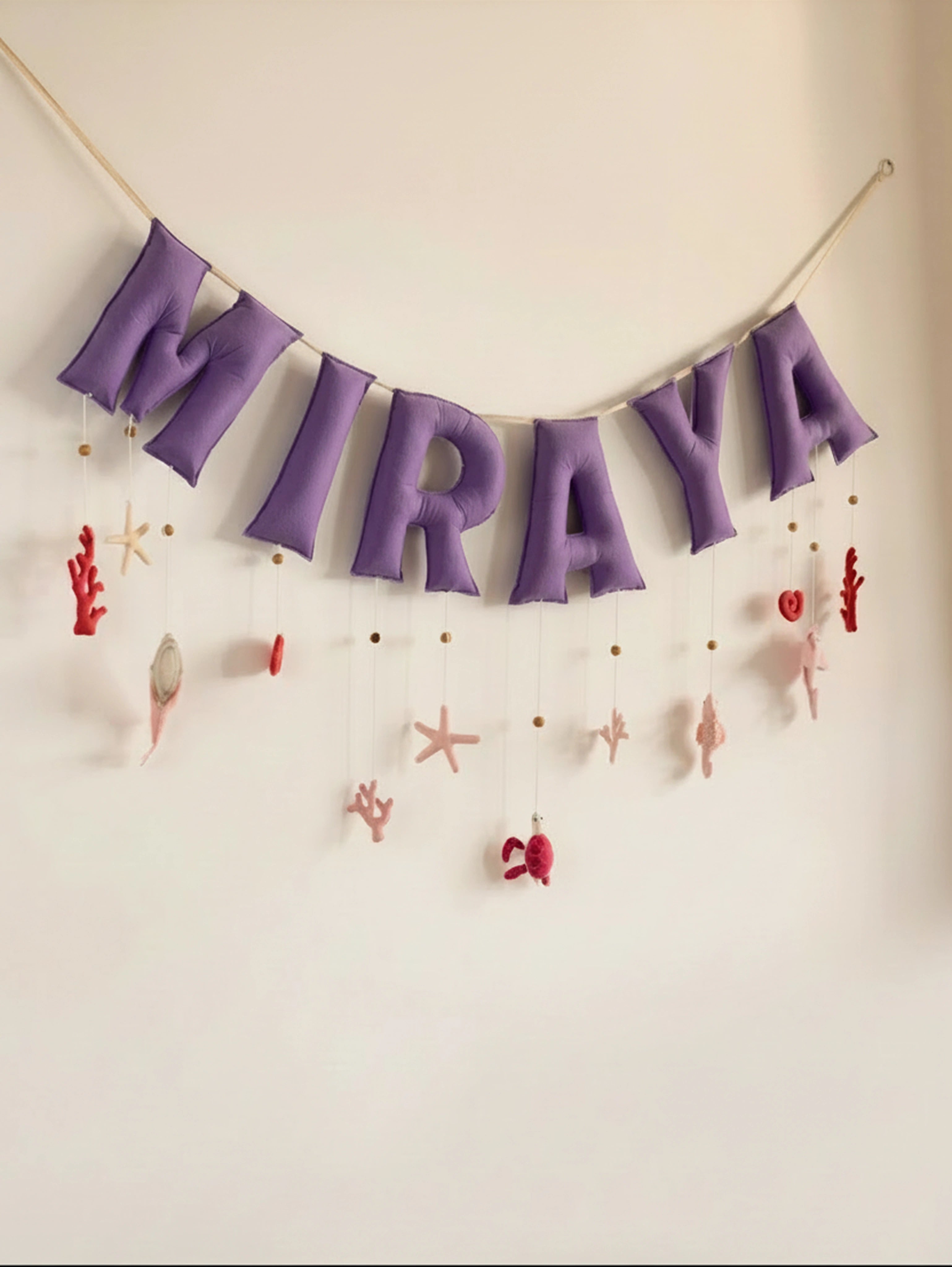 Personalized Miraya felt name banner with purple letters and handcrafted ocean-themed nursery decorations, including seashells, coral, and starfish