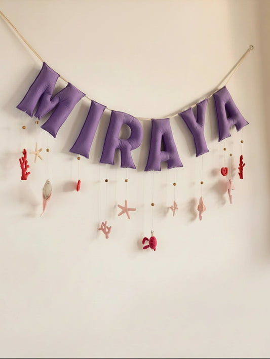 Personalized Miraya felt name banner with purple letters and handcrafted ocean-themed nursery decorations, including seashells, coral, and starfish